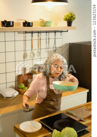 Senior woman smelling food in a frying pan while cooking in the kitchen Senior woman smelling food in a frying pan while cooking in the kitchen 123823154