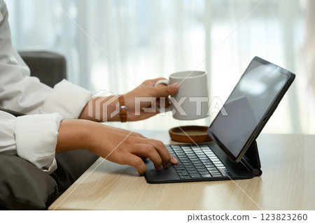 Close up of hands typing on digital tablet keyboard, holding a coffee mug Close up of hands typing on digital tablet keyboard, holding a coffee mug 123823260