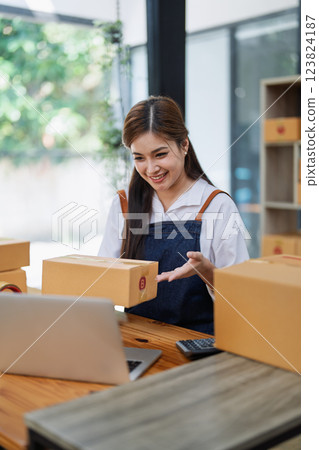 Young woman engaged in online business while packing delivery boxes at a wooden table while working on laptop for video to customer, Online selling and logistics concepts Young woman engaged in online business while packing delivery boxes at a wooden table while working on laptop for video to customer, Online selling and logistics concepts 123824187