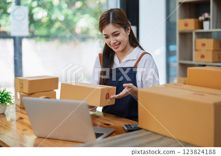 Young woman engaged in online business while packing delivery boxes at a wooden table while working on laptop for video to customer, Online selling and logistics concepts Young woman engaged in online business while packing delivery boxes at a wooden table while working on laptop for video to customer, Online selling and logistics concepts 123824188