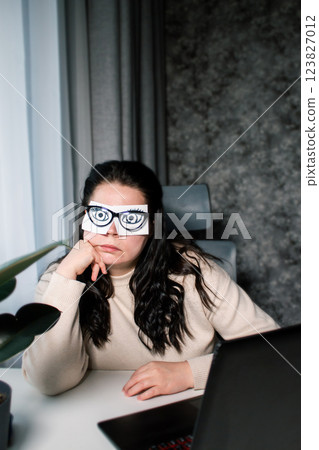Overworking at office concept. No time to sleep. Female office worker with painted eyes on stickers imitating working in front of computer while sleeping sitting at desk Overworking at office concept. No time to sleep. Female office worker with painted eyes on stickers imitating working in front of computer while sleeping sitting at desk 123827012