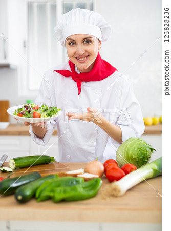 Smiling female chef in white uniform offering plate of vegetable salad in home kitche Smiling female chef in white uniform offering plate of vegetable salad in home kitche 123828236