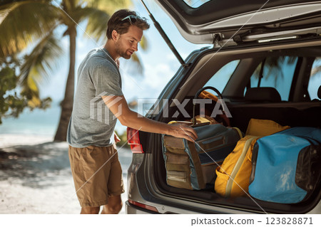 Smiling man in casual wear unpacking colorful luggage from the trunk of his car under lush palm trees on a sunny day 123828871