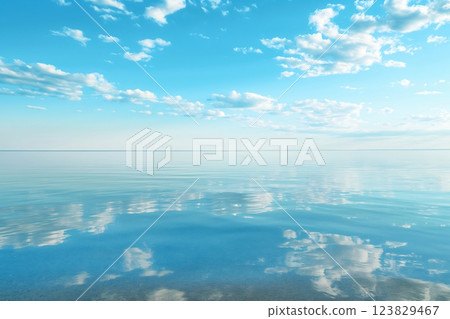 Tranquil beach scene with calm, crystal-clear water reflecting a beautiful sky dotted with fluffy clouds at the horizon 123829467