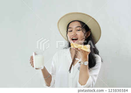 Young Asian woman in straw hat passionately eating bread in her hand and in her hand holding a glass of milk, woman breakfast concept, isolated white background Young Asian woman in straw hat passionately eating bread in her hand and in her hand holding a glass of milk, woman breakfast concept, isolated white background 123830384