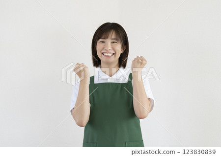 A woman in her 30s wearing an apron smiling and making a fist pump, looking at the camera A woman in her 30s wearing an apron smiling and making a fist pump, looking at the camera 123830839