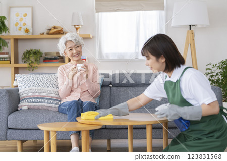 家事服務圖片：一位 30 多歲的女性正在打掃房間，一位老年女性在沙發上休息 123831568
