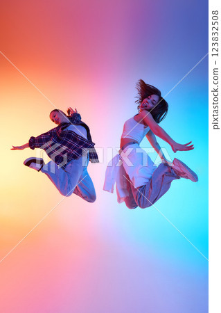 Two energetic young women showcasing bold hip hop moves against vibrant neon gradient studio backdrop. Two energetic young women showcasing bold hip hop moves against vibrant neon gradient studio backdrop. 123832508