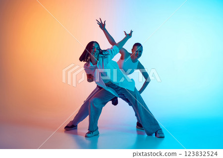 Energetic young women in jeans performing hip hop moves against gradient studio background in neon light. Energetic young women in jeans performing hip hop moves against gradient studio background in neon light. 123832524