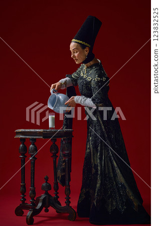 Elderly woman in renaissance clothing carefully pouring milk from ceramic jug into glass, evoking classical still life paintings against deep red background. 123832525