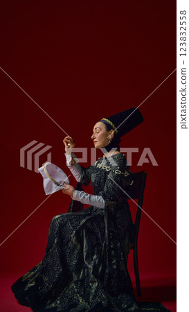 Elderly woman in historical noble dress carefully embroidering fabric with concentrated expression, hands gracefully working against deep red background. 123832558
