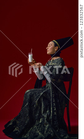 Elderly woman in renaissance-style dress gracefully holding glass of milk with striped straw, gazing upwards with serene expression against deep red background. Elderly woman in renaissance-style dress gracefully holding glass of milk with striped straw, gazing upwards with serene expression against deep red background. 123832567