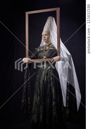 Young noblewoman in renaissance dress holding wooden frame around her upper body, looking forward with confident expression against dark background. Young noblewoman in renaissance dress holding wooden frame around her upper body, looking forward with confident expression against dark background. 123832586