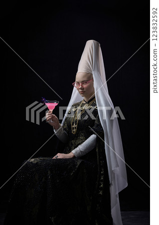 Young noblewoman in renaissance dress wearing pink sunglasses, holding martini glass with bright pink drink, gazing confidently against dark background. Young noblewoman in renaissance dress wearing pink sunglasses, holding martini glass with bright pink drink, gazing confidently against dark background. 123832592