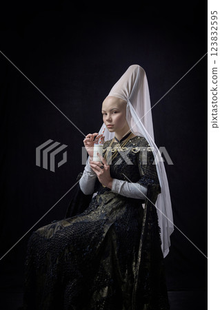 Young noblewoman in renaissance dress holding glass of milk with straw, gazing forward with calm look against dark background. 123832595