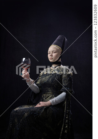 Young confident woman dressed renaissance style clothes, sitting with calm expression, holding glass of red wine against dark studio background. 123832609