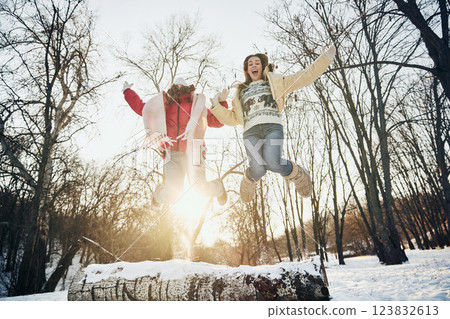 Two friends, women, dressed cozy and warm winter clothes, jumping excitedly in snowy park, arms raised in happiness. Two friends, women, dressed cozy and warm winter clothes, jumping excitedly in snowy park, arms raised in happiness. 123832613