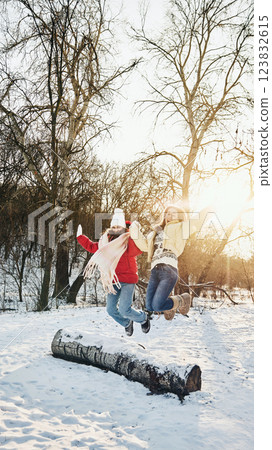Two friends in warm jackets jumping over snowy log, enjoying outdoor adventure and having fun in snowy park. 123832615