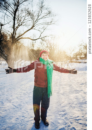 Joyful young man dressed in red hat and green scarf throws hands up, smiling while snowflakes fall in sunny park. 123832628