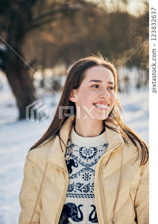 Cheerful young woman in beige winter coat and patterned sweater looks away with happiness on sunny day in park. Cheerful young woman in beige winter coat and patterned sweater looks away with happiness on sunny day in park. 123832637