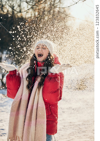 Young woman in red puffer jacket and white hat throws snow in air, laughing with joy and happiness on sunny winter day in park. 123832645
