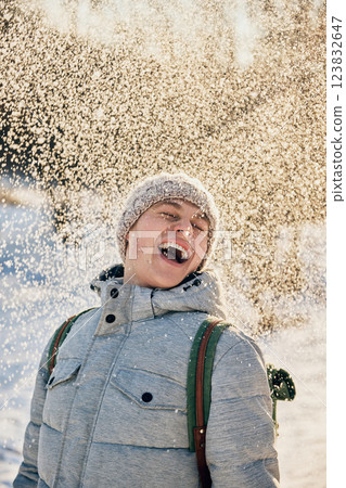 Smiling young man dressed in gray winter jacket and knitted hat enjoys snowfall in sunny winter park. 123832647