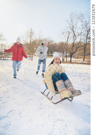 Excited young woman riding sled, being chased by laughing friends, enjoying thrilling winter adventure. 123832679