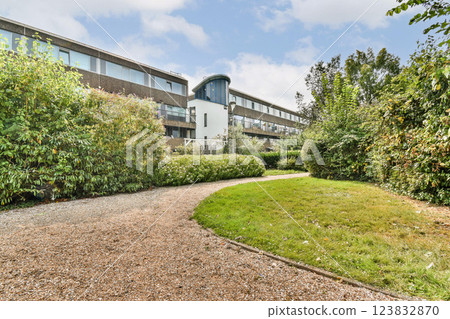 A winding pathway leads through vibrant greenery and well-maintained gardens adjacent to a modern building, embodying tranquility and natural beauty. 123832870
