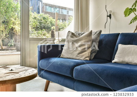 A stylish living room featuring a blue sofa, textured cushions, and large windows that open to a lush garden, creating a perfect blend of comfort and nature. A stylish living room featuring a blue sofa, textured cushions, and large windows that open to a lush garden, creating a perfect blend of comfort and nature. 123832933
