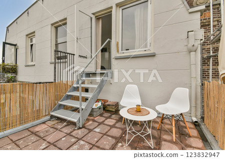 A modern outdoor patio featuring a staircase, two white chairs, and a small table on a tiled surface, surrounded by a wooden fence. 123832947