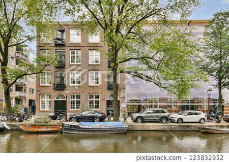 A picturesque view of a classic Amsterdam building alongside a tranquil canal, surrounded by lush green trees and parked boats, reflecting the city's unique architecture. 123832952