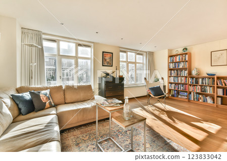 A spacious living room featuring large windows, a comfy sofa, and a rich variety of books on the shelves, creating a warm and inviting atmosphere. 123833042