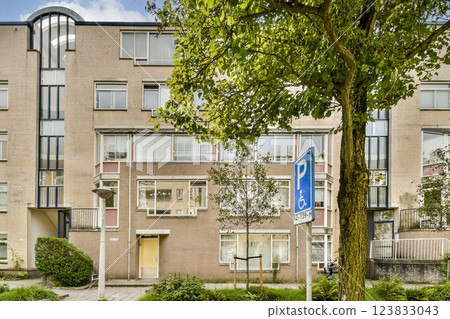 A contemporary residential building featuring large windows and a green exterior. The surrounding area includes trees and a parking sign, enhancing the urban environment. 123833043