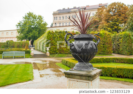 Classical stone urn on carved pedestal with ornate details. Garden with stone urn and plants. Outdoor garden with urn and flowers. Landscape with stone planter and greenery. Garden scene with 123833129