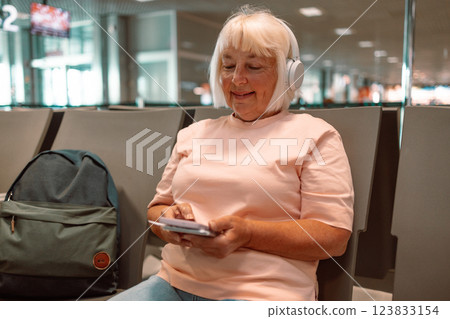Elderly woman use smartphone and earphones while waiting for her flight, 50s 60s Lady girl listening to music or podcast at the airport while waiting for a flight Elderly woman use smartphone and earphones while waiting for her flight, 50s 60s Lady girl listening to music or podcast at the airport while waiting for a flight 123833154