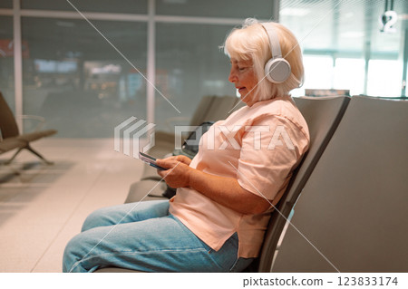 Elderly woman use smartphone and earphones while waiting for her flight, 50s 60s Lady girl listening to music or podcast at the airport while waiting for a flight 123833174