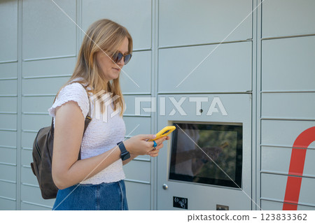 Cheerful young travel 30s woman using smart phone while standing near automatic lockers. New technologies in delivery service, self picking Cheerful young travel 30s woman using smart phone while standing near automatic lockers. New technologies in delivery service, self picking 123833362
