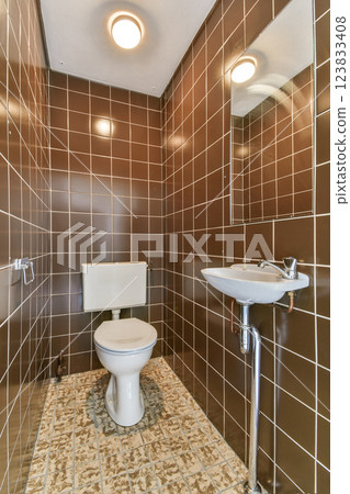 A contemporary bathroom featuring brown ceramic tiles, a white toilet, and a small sink. The design emphasizes simplicity and functionality. 123833408