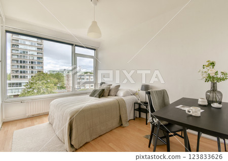 A bright modern bedroom showcasing a neatly made bed, stylish furniture, and large windows that offer a glimpse of the cityscape outside. A bright modern bedroom showcasing a neatly made bed, stylish furniture, and large windows that offer a glimpse of the cityscape outside. 123833626