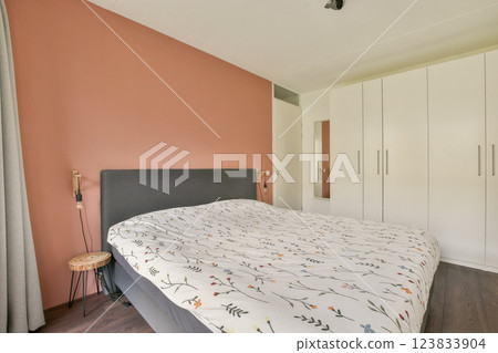 A modern bedroom featuring a neatly made bed with floral bedding, minimalistic decor, and a calming color palette. The wardrobe complements the serene atmosphere. 123833904