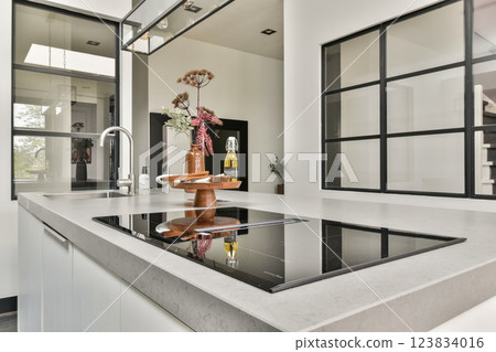 A stylish modern kitchen featuring a minimalist design with a centrally positioned induction cooktop and elegant decor elements including plants and accessories. 123834016