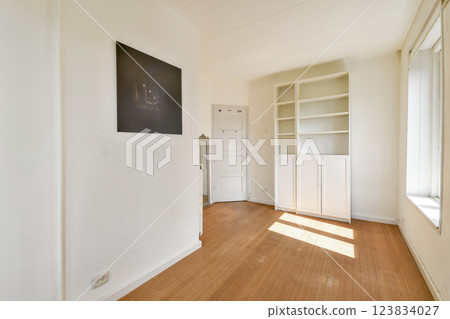 A bright and minimalist room featuring light hardwood flooring, a built-in shelf, and large windows letting in sunlight, creating a serene atmosphere. 123834027