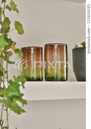 Chic gradient glass cups are arranged on a shelf adorned with lush green plants, creating a stylish and refreshing atmosphere in the space. Chic gradient glass cups are arranged on a shelf adorned with lush green plants, creating a stylish and refreshing atmosphere in the space. 123834283