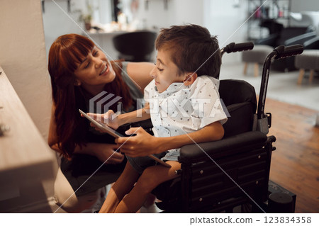 Woman, child with a disability and tablet in wheelchair friendly salon for and inclusivity, special needs and hairdressing support. Kid with cerebral palsy, screen time and games in hairdresser shop. Woman, child with a disability and tablet in wheelchair friendly salon for and inclusivity, special needs and hairdressing support. Kid with cerebral palsy, screen time and games in hairdresser shop. 123834358
