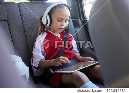 Child with tablet, headphones and safety belt in the car going home after soccer match, game or practice. Young girl in sports gear playing educational games online on tech device after training 123834381