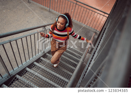 City, music and girl on stairs, phone in hand and listening to audio. Happy black woman with headphones streaming song, track or radio on smartphone in urban town, student freedom on weekend City, music and girl on stairs, phone in hand and listening to audio. Happy black woman with headphones streaming song, track or radio on smartphone in urban town, student freedom on weekend 123834385