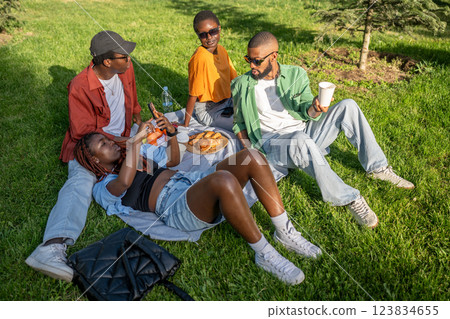 African American friends picnic in park, lying on grass with smartphones, sunbathing and snacking 123834655