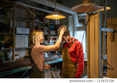 Smiling female carpenter humorously shakes sawdust off husband head in workshop. Break on work Smiling female carpenter humorously shakes sawdust off husband head in workshop. Break on work 123835106