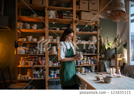 Female potter hold and attach handle to clay mug for material bonding, preparing for drying, firing Female potter hold and attach handle to clay mug for material bonding, preparing for drying, firing 123835115