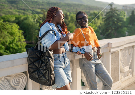Laughing black friends chatting while lean on railing, waiting for taxi after booking on smartphone Laughing black friends chatting while lean on railing, waiting for taxi after booking on smartphone 123835131